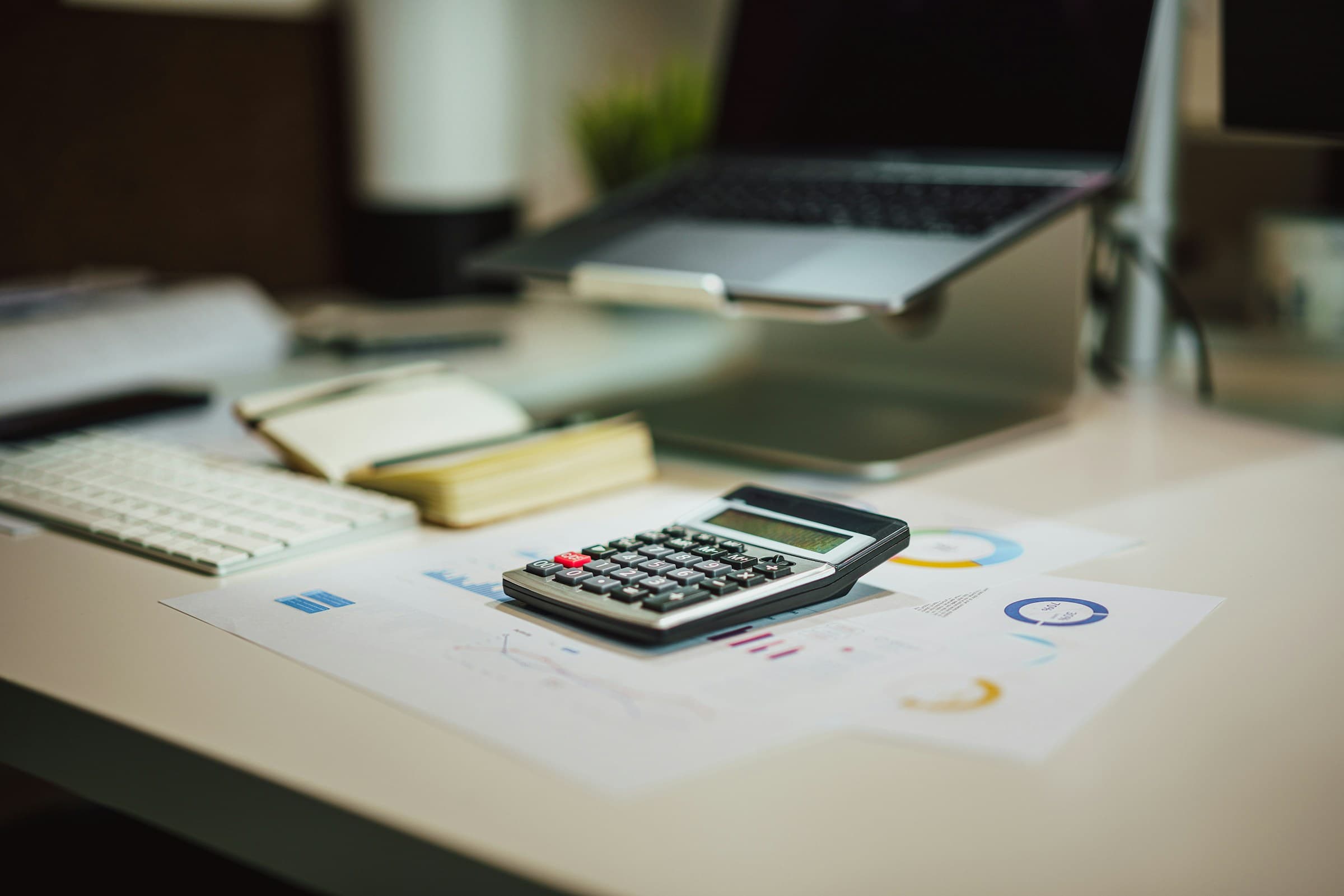 A desk with a calculator on printed financial charts, a notebook, a keyboard and a laptop.